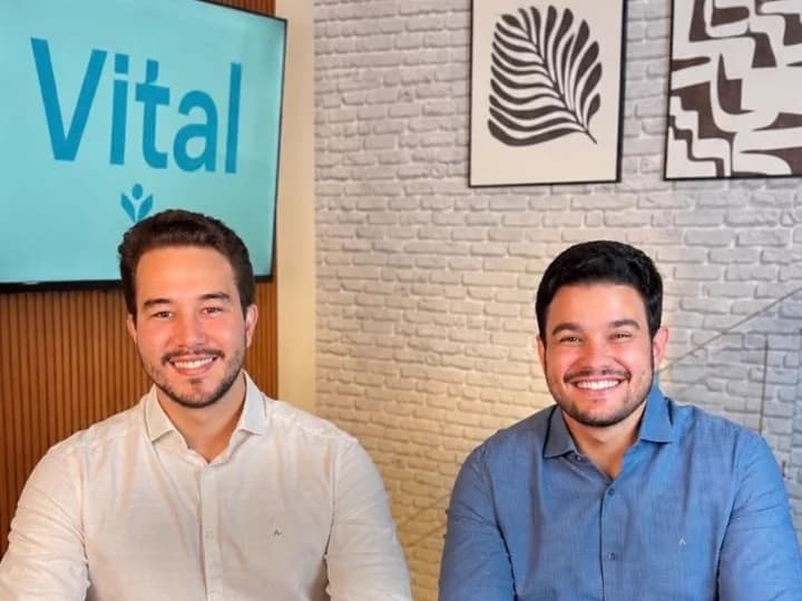 Os dois sócios fundadores da Vital sorrindo, sentados lado a lado e olhando para a câmera, com o logo da Vital numa TV ao fundo